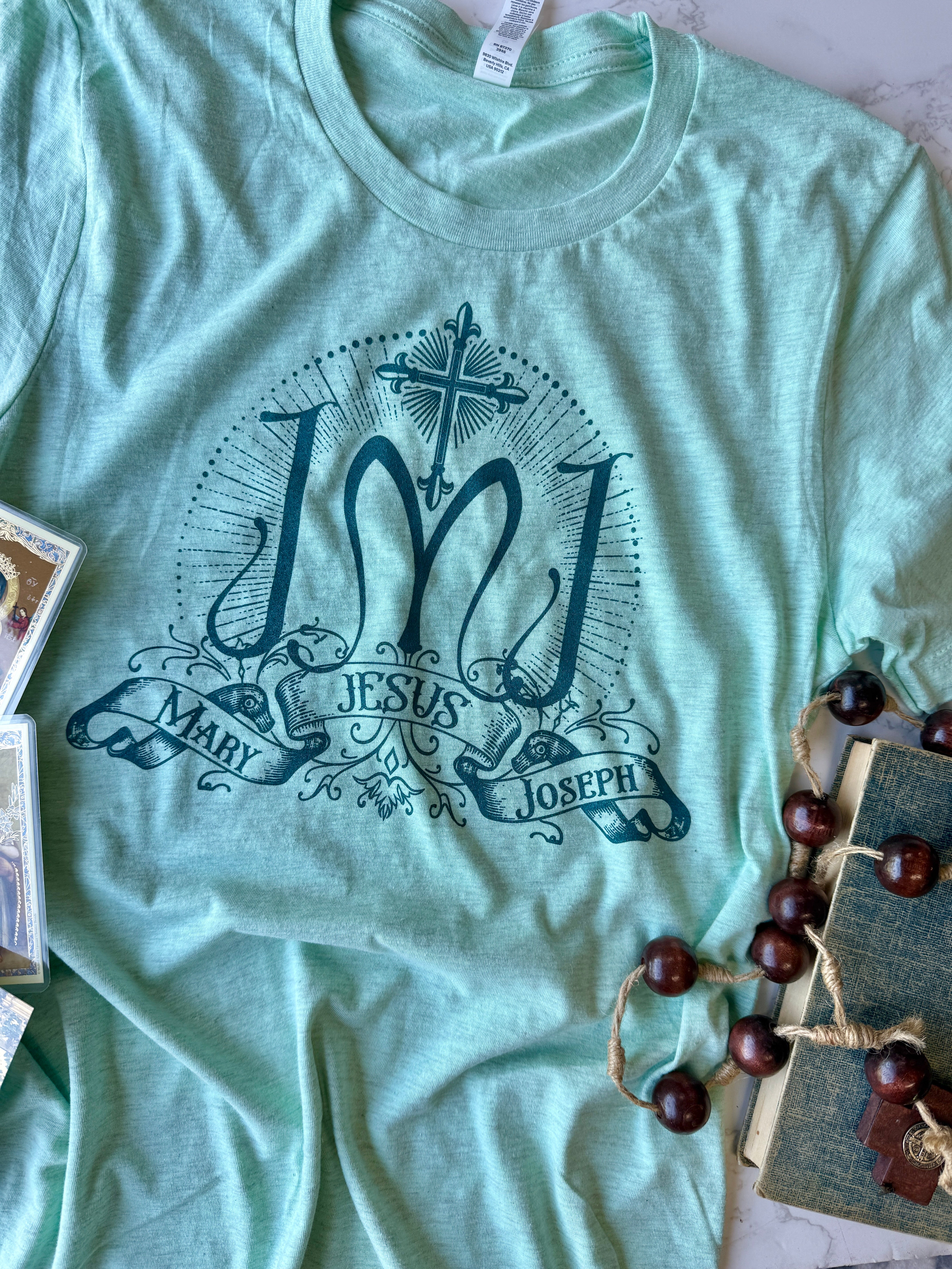 Light green t-shirt with 'Jesus Mary Joseph' design on a surface with decorative items.