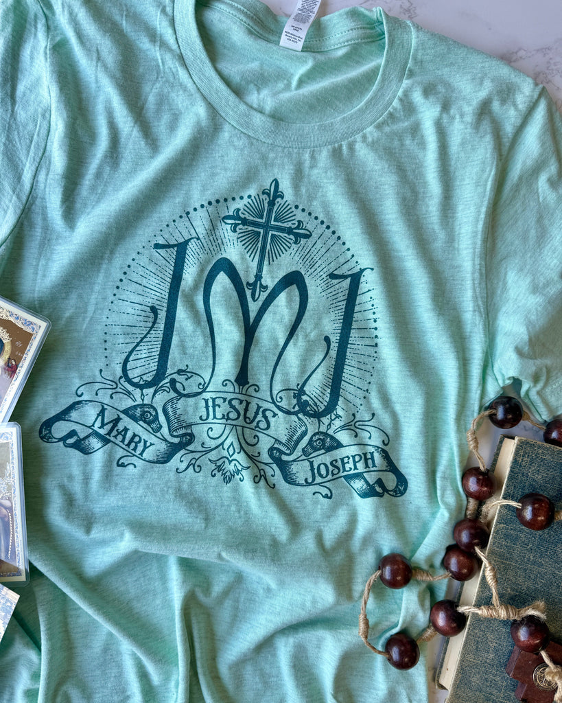 Light green t-shirt with 'Jesus Mary Joseph' design on a surface with decorative items.